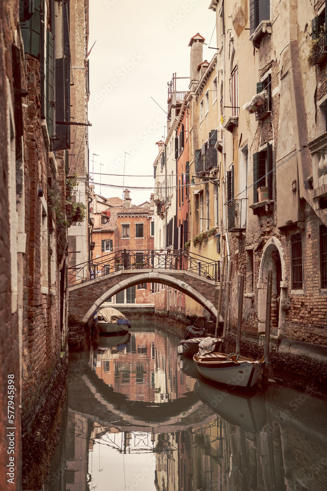 Arched bridge over a narrow canal in Venice with old houses and parked boats next to them (vintage style photo)