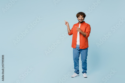 Full body young fun Indian man wear orange red shirt white t-shirt point index finger aside indicate on workspace area copy space mock up isolated on plain pastel blue cyan background studio portrait.