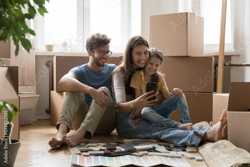 Positive parents and happy kids taking moving selfie on smartphone, sitting at stacked cardboard boxes, renovation brochures with wall color samples, holding mobile phone, smiling
