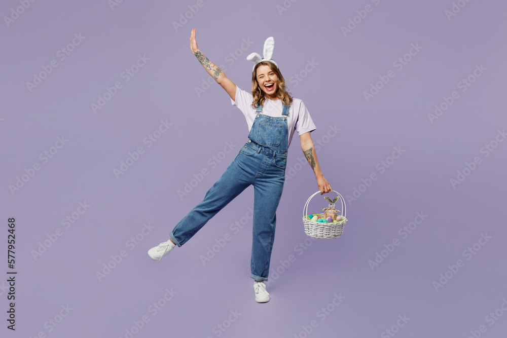 Full body smiling fun young woman wearing casual clothes bunny rabbit ears holding wicker basket colorful eggs raising hand up isolated on plain pastel purple background studio. Happy Easter concept. - obrazy, fototapety, plakaty 