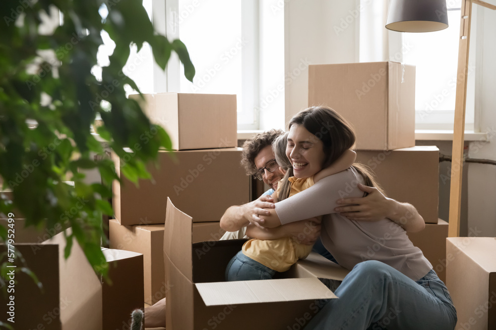 Happy joyful kid sticking out cardboard moving box, hugging mom and dad. Cheerful couple of parents playing with daughter girl among relocation containers, unpacking stuff at new home Happy joyful kid sticking out cardboard moving box, hugging mom and dad. Cheerful couple of parents playing with daughter girl among relocation containers, unpacking stuff at new home
