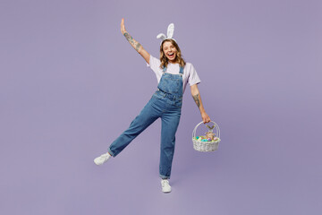 Full body smiling fun young woman wearing casual clothes bunny rabbit ears holding wicker basket colorful eggs raising hand up isolated on plain pastel purple background studio. Happy Easter concept.