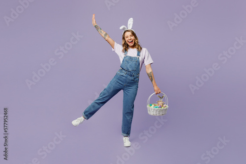 Foto Full body smiling fun young woman wearing casual clothes bunny rabbit ears holding wicker basket colorful eggs raising hand up isolated on plain pastel purple background studio
