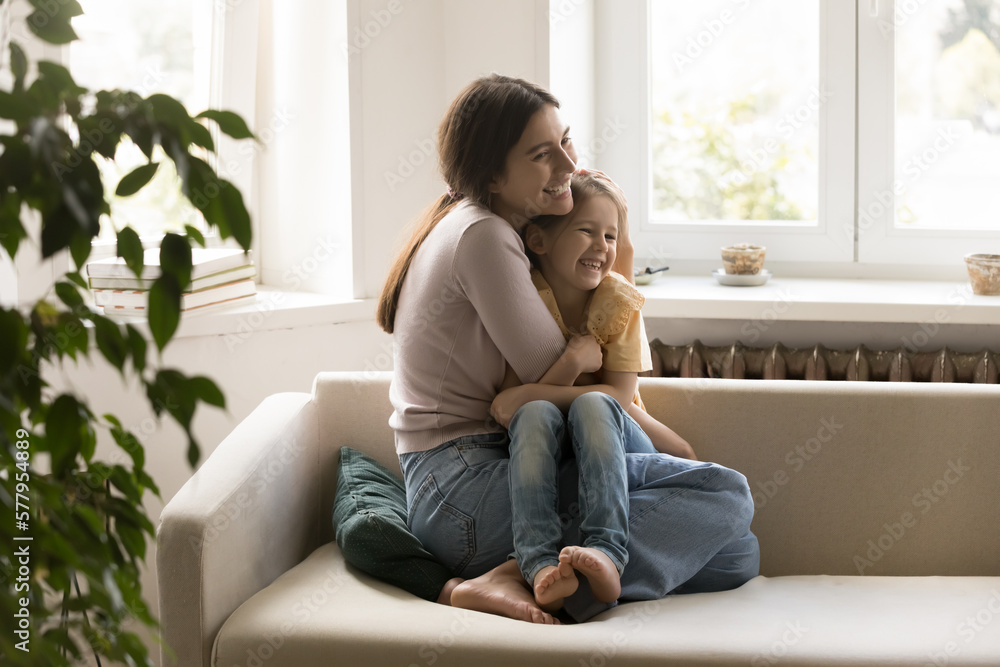 Joyful excited young mom cuddling sweet little girl on home sofa, hugging, tightening daughter kid with love, affection, tenderness, holding child in arms on couch, laughing, enjoying motherhood Joyful excited young mom cuddling sweet little girl on home sofa, hugging, tightening daughter kid with love, affection, tenderness, holding child in arms on couch, laughing, enjoying motherhood