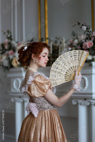 Young red haired woman in ball historic dress and fan in hand in vintage room full of flowers. Portrait of aristicratic woman in historically decorated room. photo