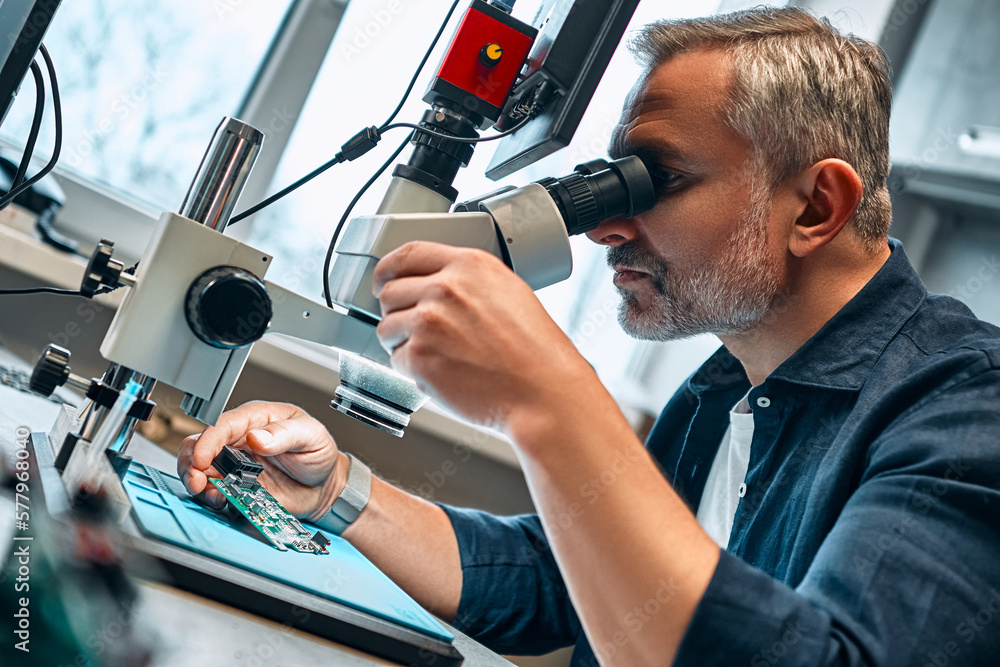 A mature man looks at a microcircuit through a microscope. Concept of engineering, chip development and service. A mature man looks at a microcircuit through a microscope. Concept of engineering, chip development and service.
