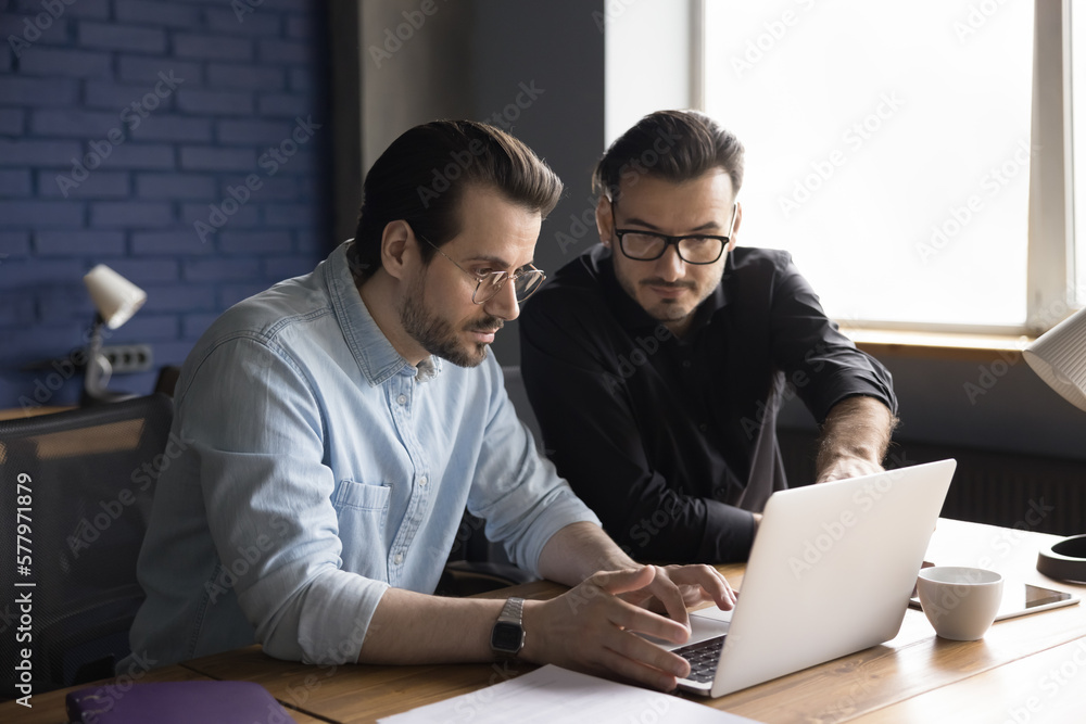 Two focused business partners men working at laptop together, looking at display, watching online presentation, cooperating on application, software product, using computer Two focused business partners men working at laptop together, looking at display, watching online presentation, cooperating on application, software product, using computer