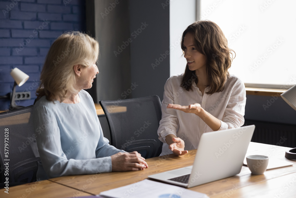 Happy positive young office professional talking to adult middle aged business woman at workplace table, discussing project at laptop with elder colleague, consulting senior mentor, teacher, expert Happy positive young office professional talking to adult middle aged business woman at workplace table, discussing project at laptop with elder colleague, consulting senior mentor, teacher, expert