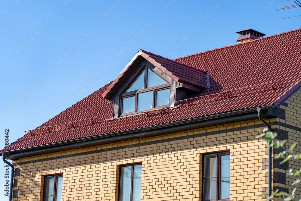 Roof of cottage made of ceramic tiles with drains, slopes, snow retainers, tides, chimney, dormer windows against blue sky. Sloping roof with metal coating of residential building with windows