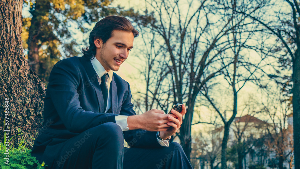 Obraz premium Happy man in a business suit sets outdoors and using a smartphone. Portrait of the young man in a business suit with mobile phone, in the park. Smiling businessman in city looking to a cell phone.