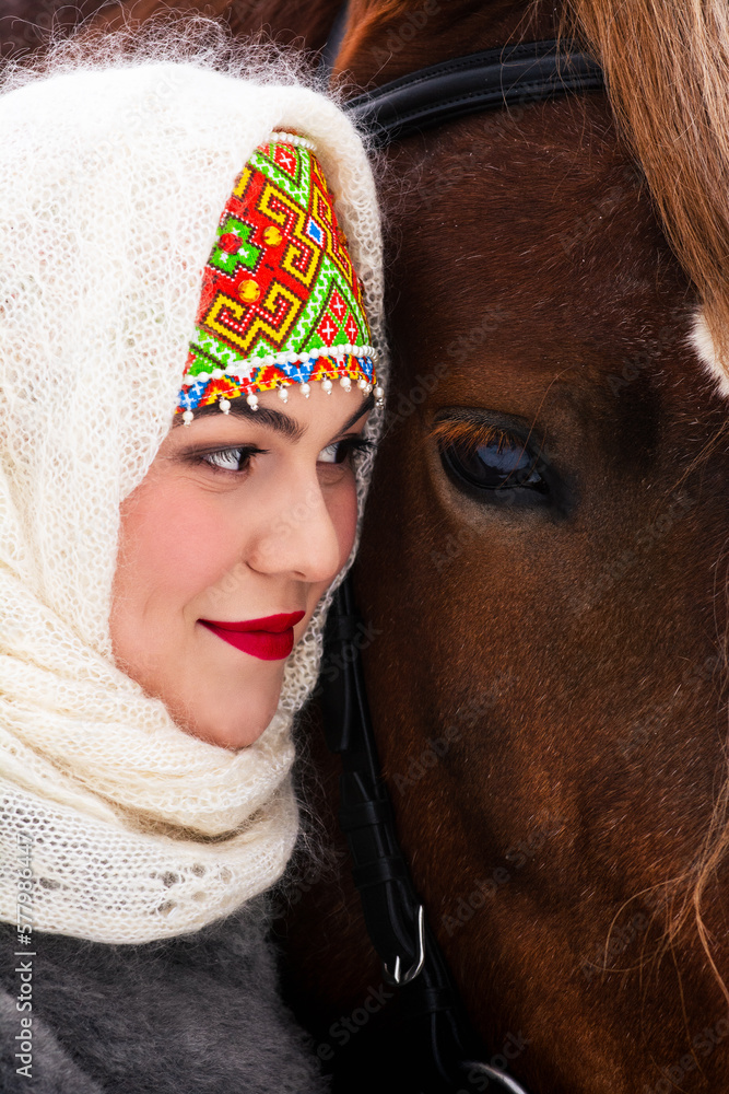 Сlose portrait of a Russian beauty girl in traditional clothes with red big horse in forest winter.