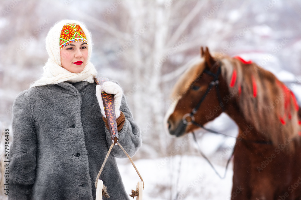Russian beauty girl in traditional clothes stands with a yoke and buckets. Behind red big horse in forest winter