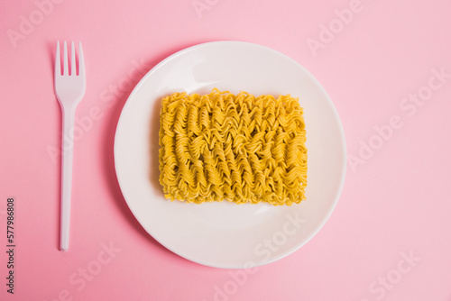 Raw instant noodles on a white plate with a plastic fork, on a pink background, flatlay. Fast food in the modern world.