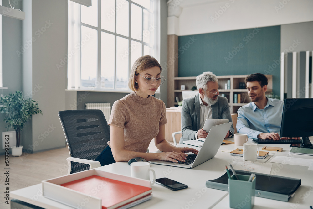 © gstockstudio - Group of confident business people working together while sitting at the shared desk in the office