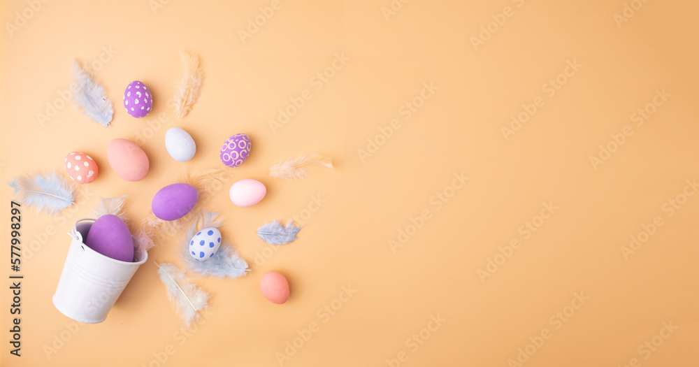 Colorful Easter eggs with feathers in decorative bucket on beige background, flat lay. Space for text