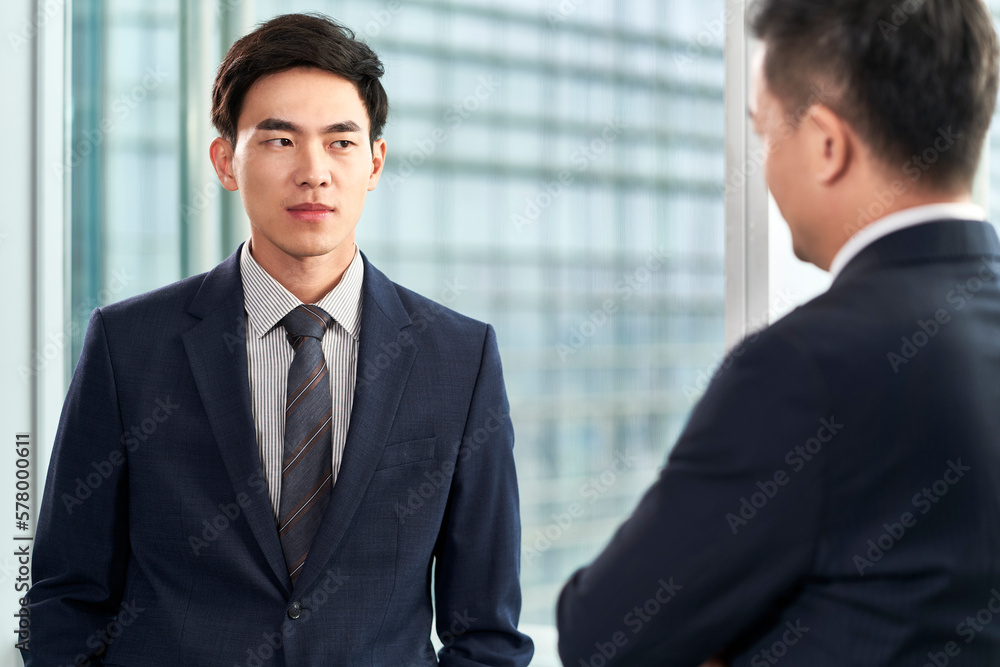 two asian businessmen standing by the window chatting talking discussing business in office two asian businessmen standing by the window chatting talking discussing business in office