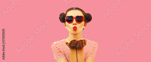 Portrait of stylish young woman blowing her lips sending sweet air kiss with lipstick wearing red heart shaped sunglasses with cool hairstyle on pink background © rohappy