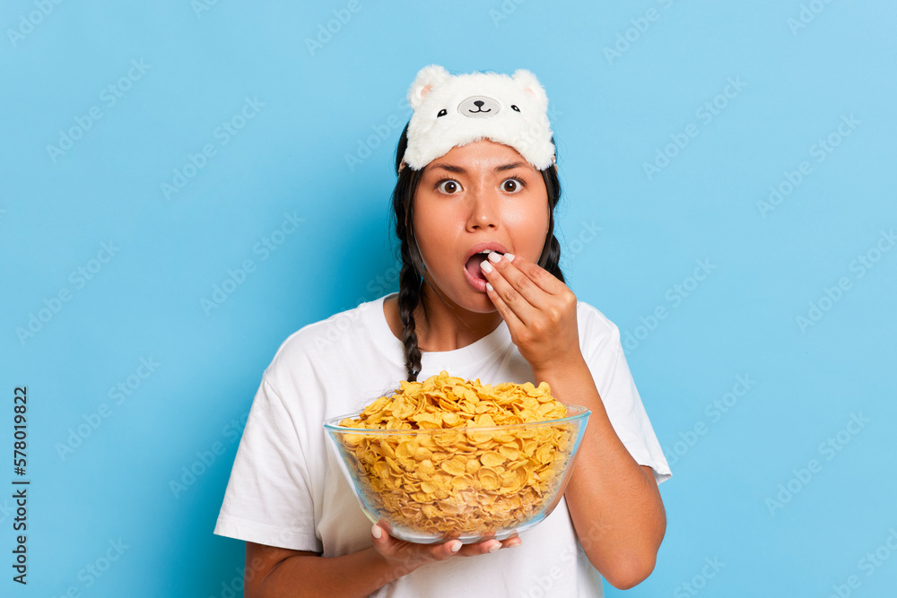 Portrait of enthusiastic asian young woman with pigtails and sleeping mask watching a tv eating corn flakes from the bowl isolated over blue studio background. People lifestyle morning routine concept Portrait of enthusiastic asian young woman with pigtails and sleeping mask watching a tv eating corn flakes from the bowl isolated over blue studio background. People lifestyle morning routine concept