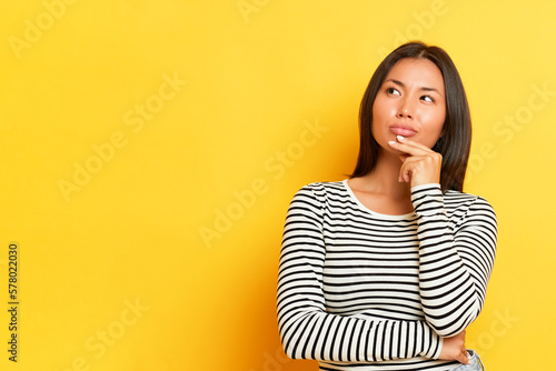 Horizontal shot of pensive Asian pretty woman keeps fingers on a chin looks thoughtfully aside blank space for promotion wears striped jumper poses over yellow background