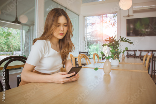 Young adult happy asian woman using smart phone for social media application, shopping online, transfer money, financial, message in cafe. Background with window and warm sunlight on summer season.