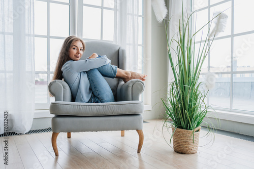 Konstfotografi Attractive blonde American young woman sitting in cozy chair hugs bended legs looks at camera smiles loves her apartments with large  windows