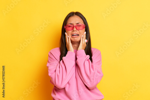 Indoor shot of hesitant brunette woman shrug shoulders put hands on the face cry and doubt while make decision stands puzzled wears modern pink sweater and pink glasses isolated over yellow background