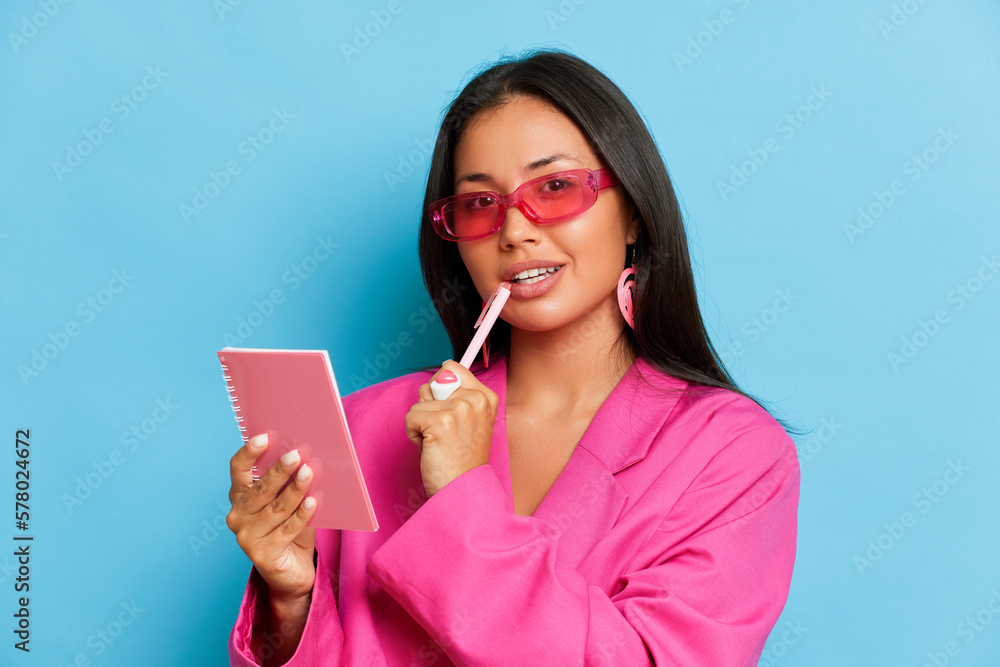 Thoughtful woman takes notes a notebook wears pink jacket and glasses isolated over blue background Thoughtful woman takes notes a notebook wears pink jacket and glasses isolated over blue background
