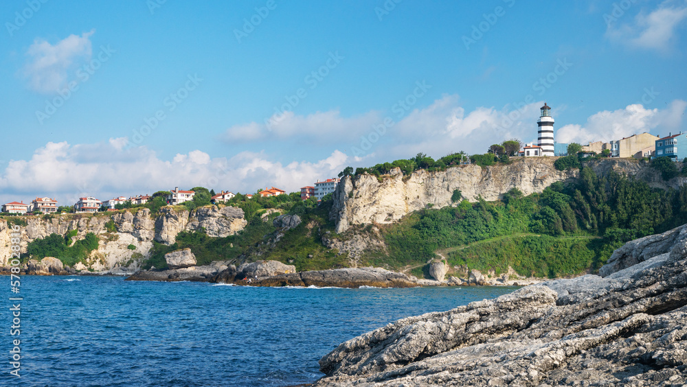 Naklejka premium A landscape with the sea and a lighthouse on a cliff with colorful houses and trees on a sunny summer day.