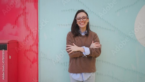 Happy woman in glasses snapping his fingers standing outdoor near colorful wall smile and talking something. Positive female fix hair laught an tel a story  photo