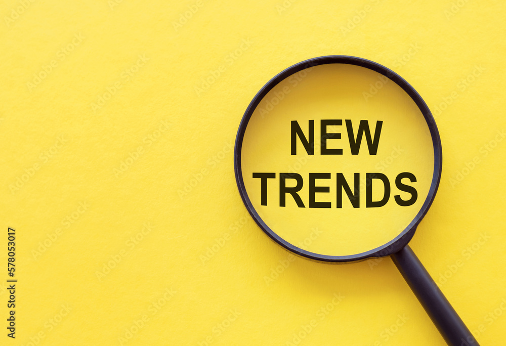 Searching new and popular trends. Magnifying glass over words on yellow background, top view Searching new and popular trends. Magnifying glass over words on yellow background, top view