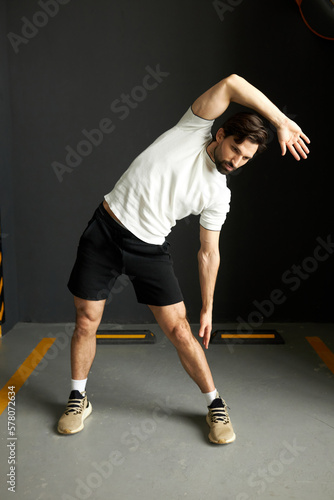 Image of muscular young sporty guy with beard stretching right hand bending to left side, training at gym on weekend, relaxing back muscles, preparing for contest, training in sport clothes