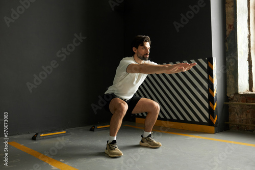 Side view indoor image of young handsome bearded fitness instructor with toned body doing squats at gym showing how to perform physical exercise right, dressed in sports clothes. Active lifestyle