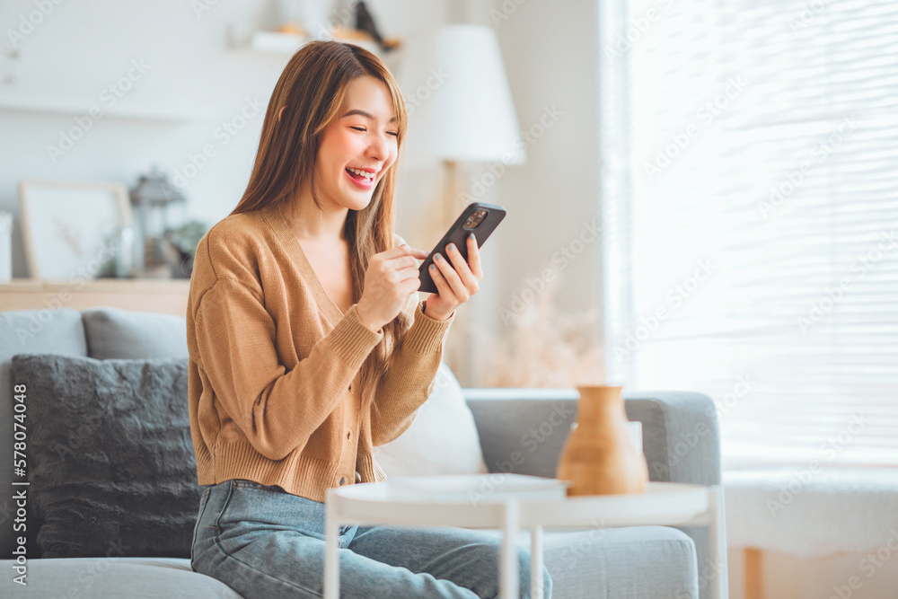 Happy young asian woman browsing surfing wireless internet on mobile phone while sitting a couch in living room at home, Shopping online via website - obrazy, fototapety, plakaty 