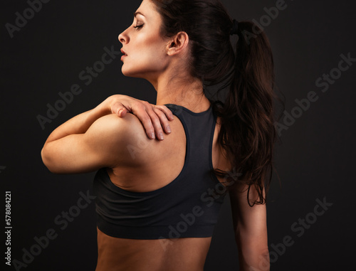 Female sporty muscular with ponytail doing stretching workout of the shoulders, blades in sport bra, holing the neck the hand on dark grey background with empty copy space. Back view. Healthy