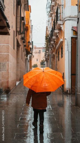 Fototapeta Naklejka Na Ścianę i Meble -  Girl with an orange umbrella protecting herself from the rain walking down a lonely narrow street