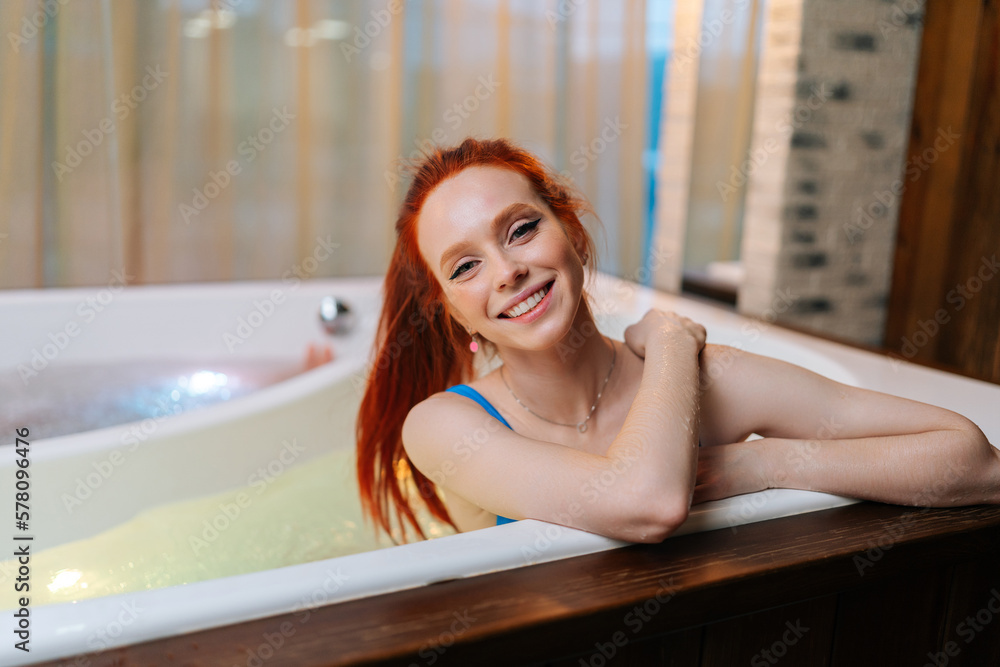 Obraz premium Portrait of charming young woman relaxed sitting in bath tub with hydromassage at luxury spa salon, smiling looking at camera. Happy female lying in bathtub relaxing hydrotherapy in beauty center.