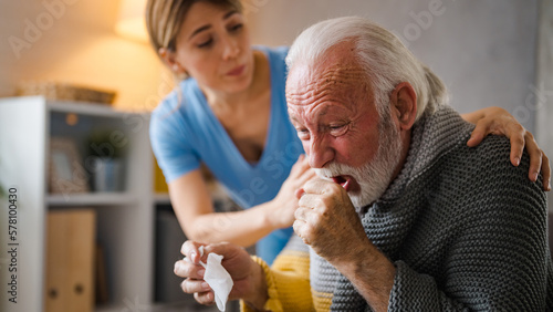 Tableau sur toile Cold exhausted senior man with flu wrapped in a warm blanket blowing his nose with a tissue in the livingroom