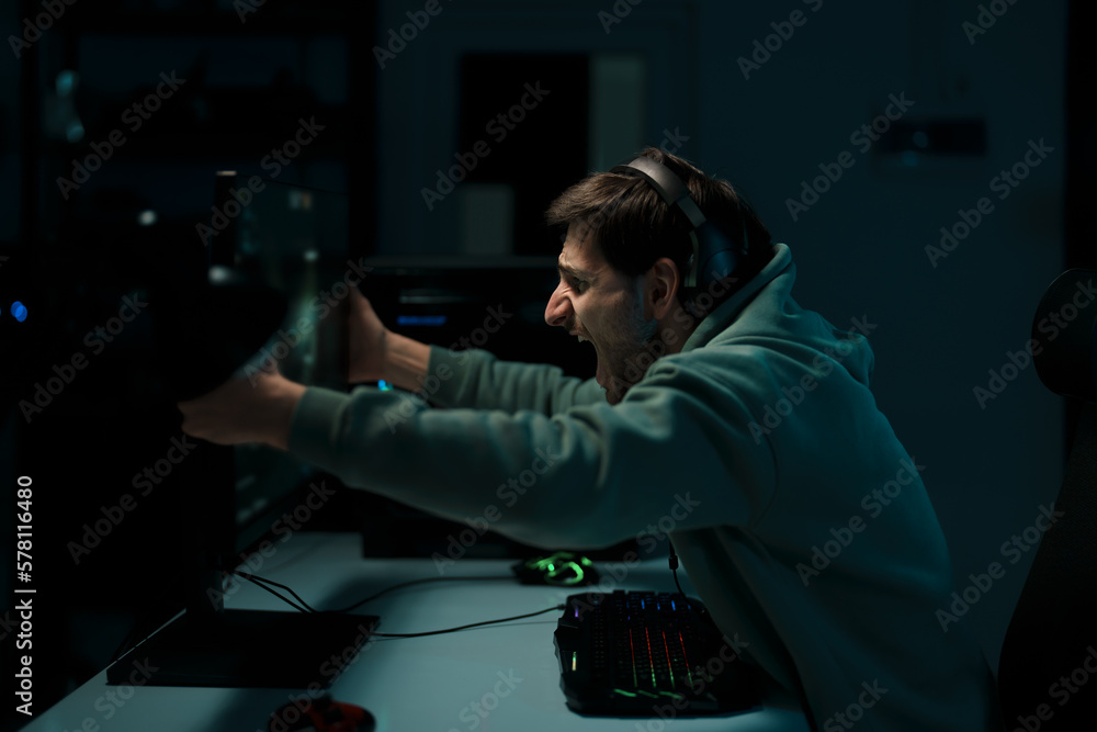 Side view of male cybersport gamer in headset touching computer monitor while celebrating successful game in dark room Side view of male cybersport gamer in headset touching computer monitor while celebrating successful game in dark room