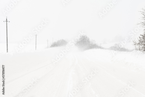 A heavy snowstorm covers the road. Large snowdrifts on the sides of a rural road. Poor visibility during a snow cyclone © Aliaksei