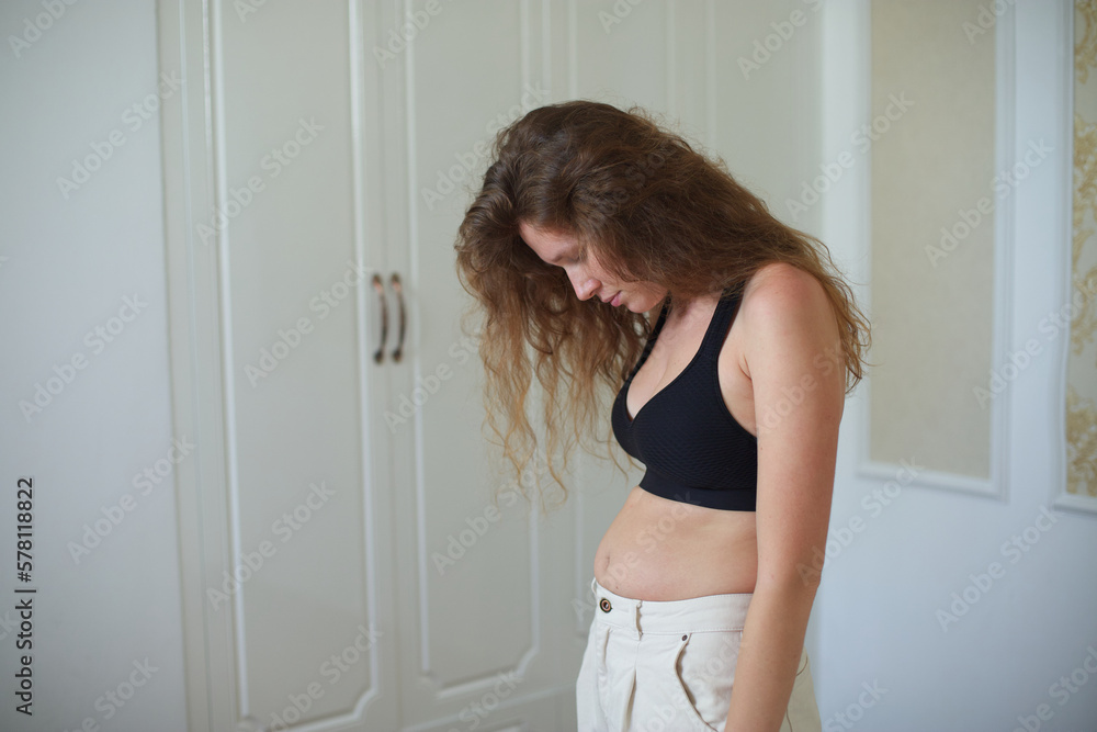 Unhappy sad upset young woman with fat on her stomach, girl worried about body fat touching her belly standing at home near closet Unhappy sad upset young woman with fat on her stomach, girl worried about body fat touching her belly standing at home near closet
