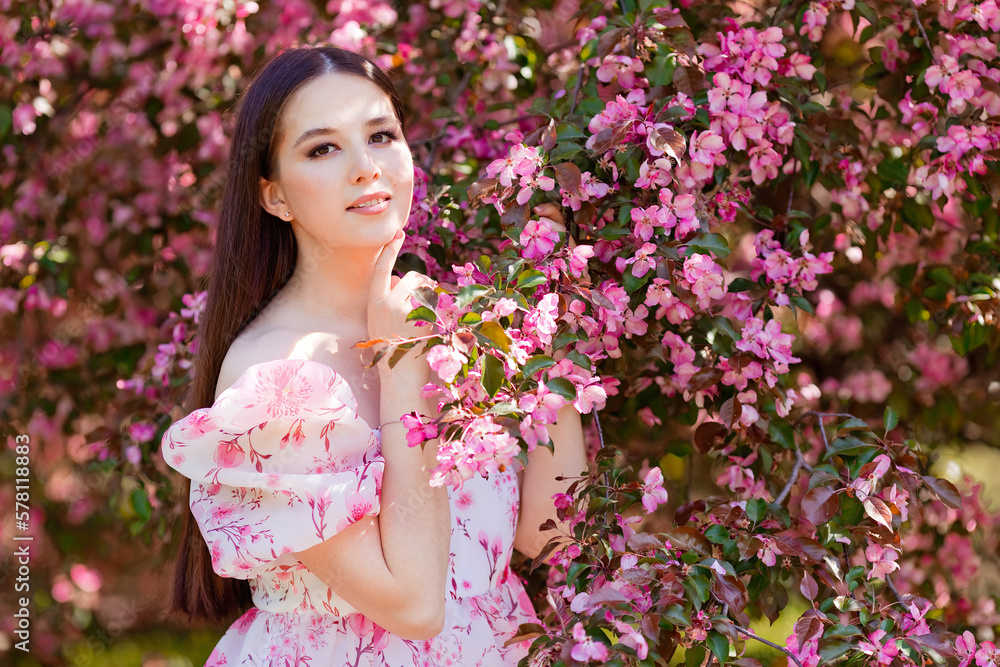 Fototapeta premium A charming girl in a pink dress standing near pink blooming garden