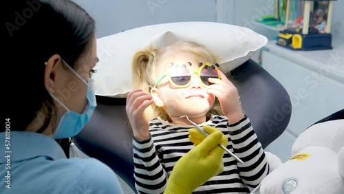 close-up face of a little girl and doctor in a mask yellow hygienic gloves dentist tools pediatric dentistry sunglasses with teddy bear eyes girl treats teeth shows doctor painless treatment photo