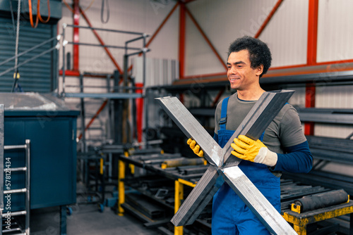 A worker wearing overalls and protective gloves holds an X-shaped metal object in a workshop