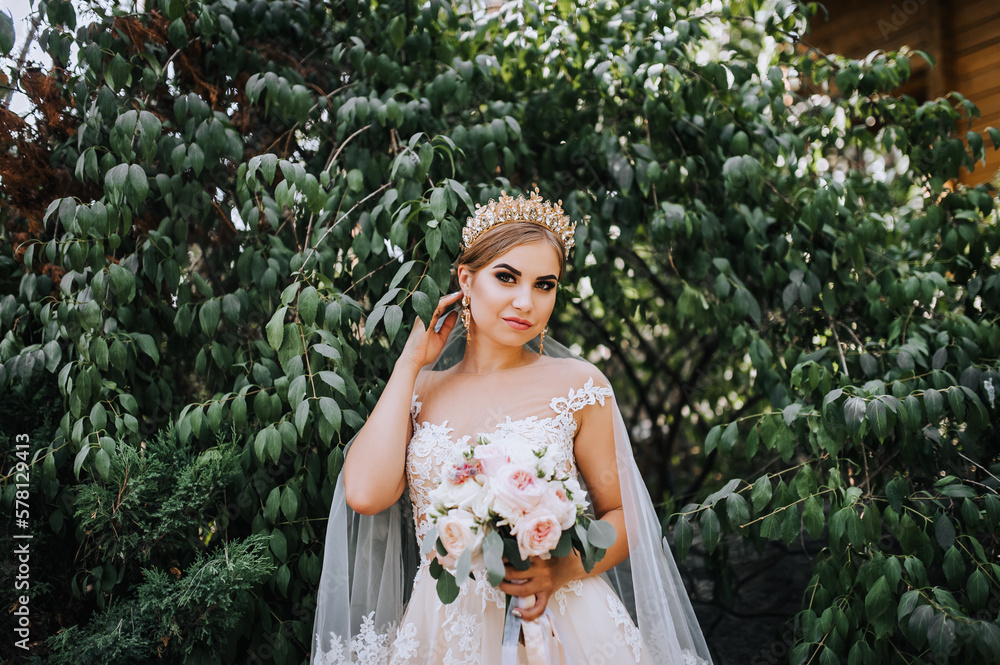 A beautiful blonde bride in a white lace dress with a crown, a diadem on her head stands in a park in nature with a bouquet in her hands. Wedding photography, portrait.
