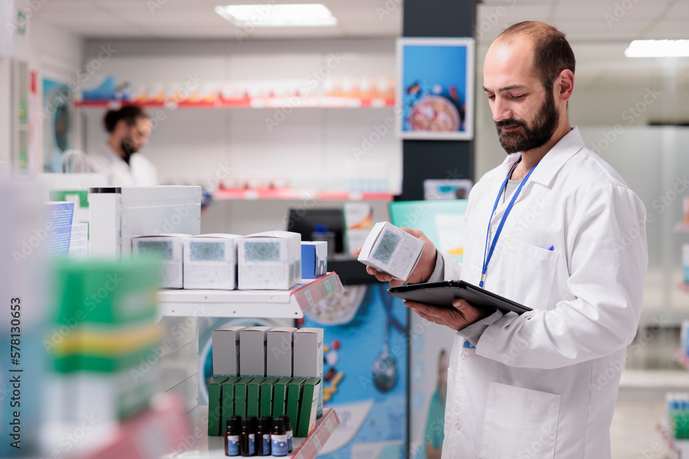 Drugstore employee looking at drugs packages typing medicaments name on tablet computer during inventory in pharmacy. Pharmacist is an expert in pharmaceuticals and is able to provide guidance
