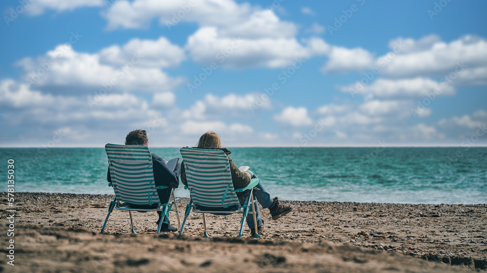 Dos personas de espaldas sentadas frente al mar leen un libro en un día soleado con cielo azul