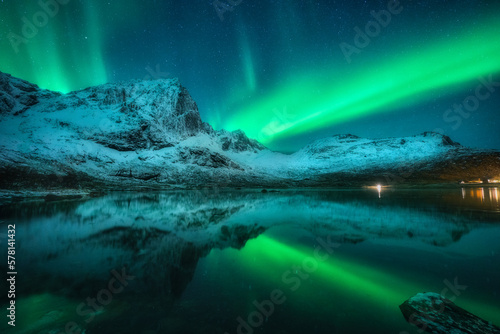 Northern lights over the snowy mountains, sea, reflection in water at night in Lofoten, Norway. Aurora borealis and snow covered rocks. Winter landscape with polar lights, sky with stars and fjord