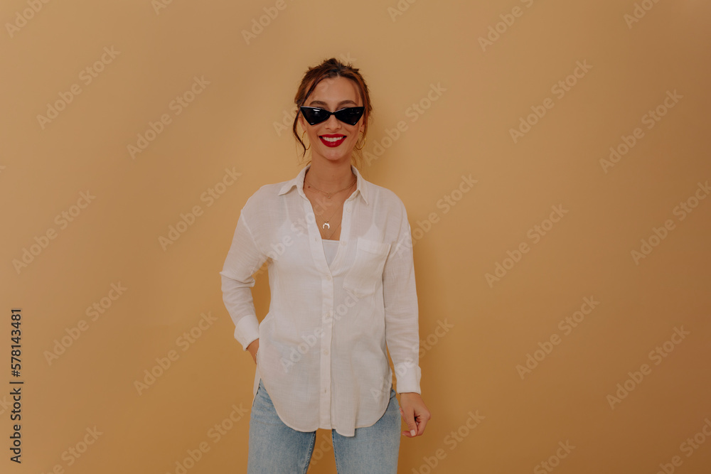 Lovely charming smiling woman with dark collected hair and red lips wearing shite shirt and jeans posing with happy smile to camera over beige background 