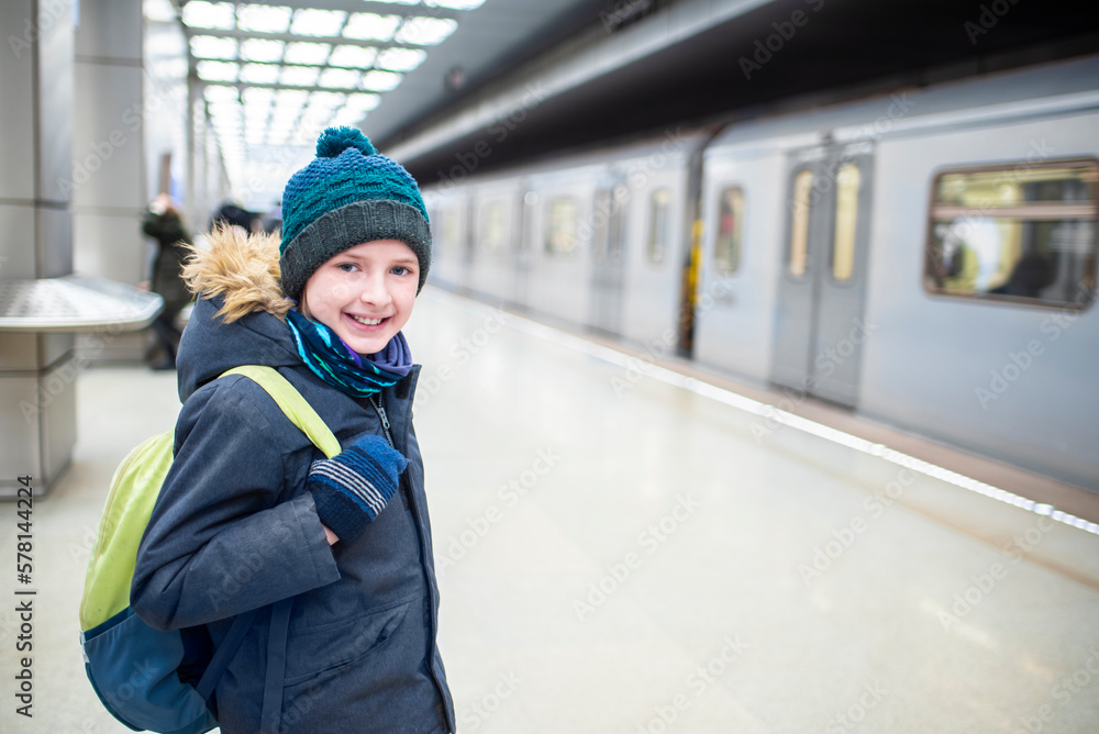a smiling boy in a winter jacket and hat is waiting for an approaching train on a subway platform