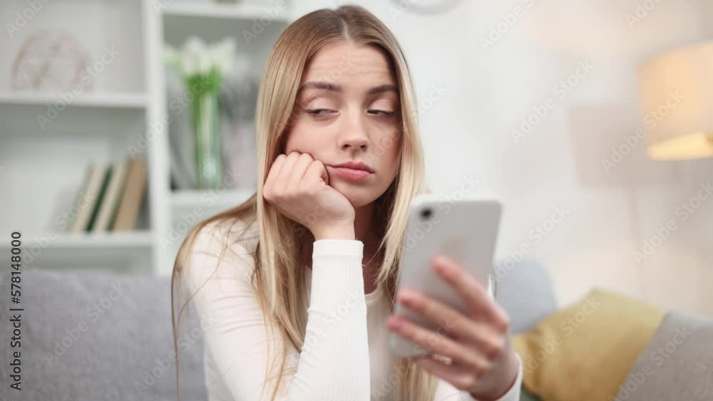 Boring young blond woman scrolling feed of social networks products in internet store on smartphone in the living room Tired exhausted female look at mobile screen without enthusiasm at home Boring young blond woman scrolling feed of social networks products in internet store on smartphone in the living room Tired exhausted female look at mobile screen without enthusiasm at home